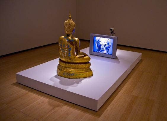 Color photo of a Nam June Paik closed circuit Buddha. Looks like a golden Thai Buddha sitting on a white platform facing a television with a camera on top that is filming him as he sits in mediation.