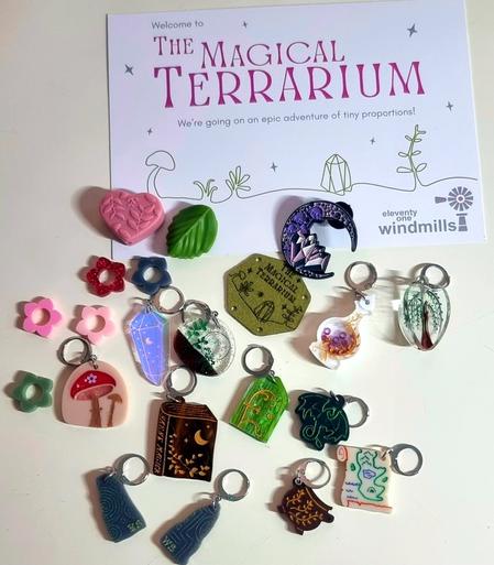 close up of 12 stitch markers from The Magical Terrarium.
designs - spellbook, crystal, mushroom, willow tree, elvish door, potion bottle, caldron, map, dragon, crystal moon, terrarium,  5 daisies. Heart and leaf needles stoppers.