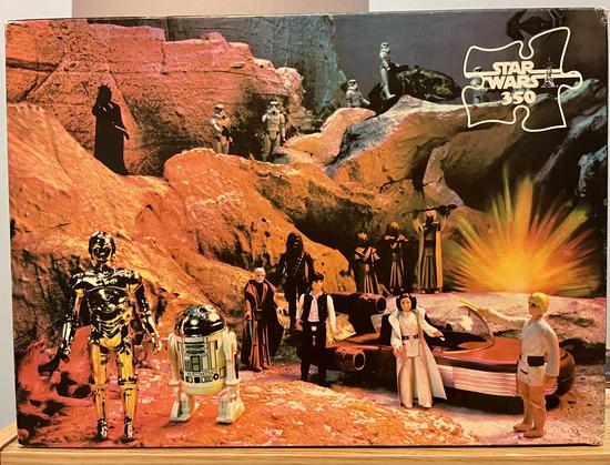 The image features a Star Wars puzzle box, showcasing various collectible figures and a scene from the franchise. Notable figures include C-3PO, R2-D2, Darth Vader, Luke, Leia, Han, and several stormtroopers, positioned around a landscape resembling Tatooine and a hover car.