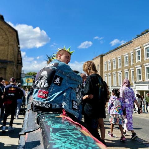 A person with a green spiked mohawk is wearing a denim jacket adorned with various patches, including a skull and crossbones. They sit on a graffiti-covered railing in a bustling street. The scene is lively, with people walking by, and the sky is clear and blue.