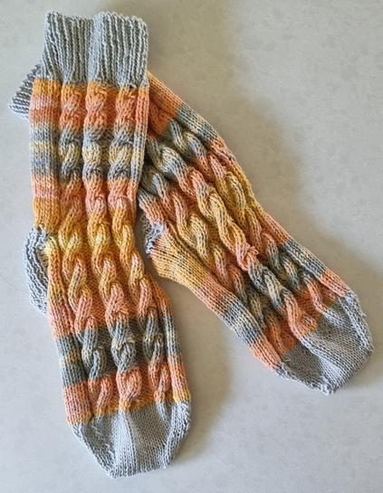 Pair of hand-kniited socks. Cuffs, heels, & toes in soft grey. Body of socks is variagated shades of yellow and oranges with bands of grey and cream
There are 3 cables running the length of each sock.