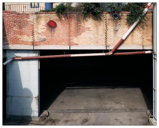 This image shows an architectural scene featuring a brick wall with varying colors and patterns on its upper section, with a partial view of a stairwell visible on the top left. There are green plants growing over the edge at the top. A large red  and white sectioned pipe runs diagonally across the wall and descends towards a garage entry. The lower section of the wall transitions into a cement structure that frames the dark entrance to the underpass or garage. The setting appears to be urban, and the composition features a blend of natural elements, utilitarian construction, and urban infrastructure. Additionally, there's a shadow of a structure or person visible on the left side, cast on the cement entryway.