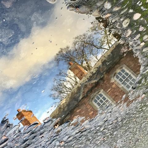 Square cropped photograph of a red clay brick building at Dunhan Massey. The building is shown in a reflection on a clean water puddle over a grey cobblestone floor. The reflection is slanted, and shows some reddish clouds. The reflection has a watercolor-like vibe.