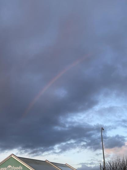 A light rainbow is set against darkened but thin and low clouds that have virga in them. There is a clearer patch of sky in the distance with light to darkening blue.