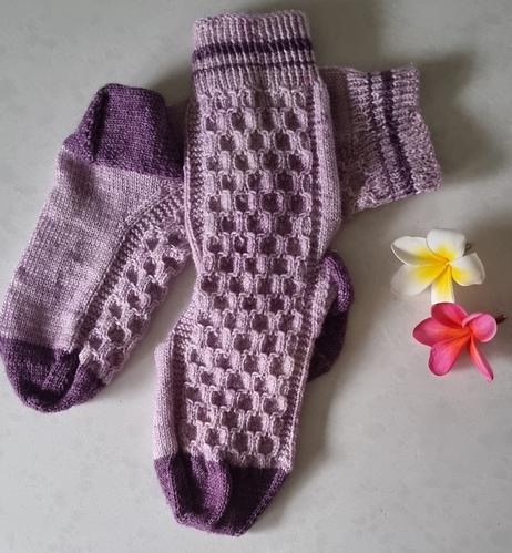Pair of hand-knitted socks. Heels, toes, are dark purple. Body of sock is mauve. Cuff is mauve with 2 bands of purple.
Pattern is like a honeycomb effect all over except for sole of foot which is done in stockinette.