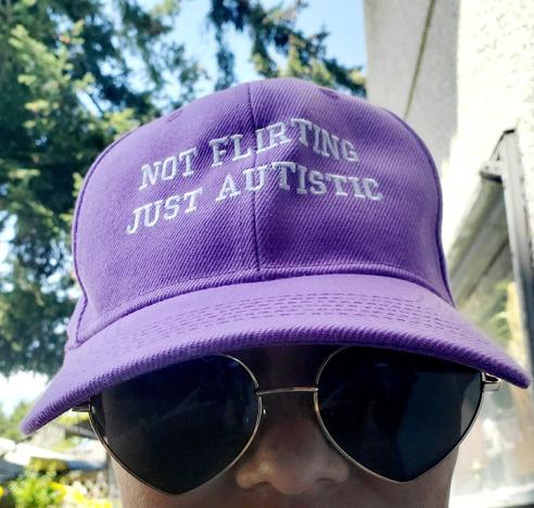 Selfie of me, wearing my bright purple baseball cap & heart-shaped sunglasses. The cap has the phrase "Not Flirting, Just Autistic" embroidered in white. The background shows a mixture of green foliage & a light-colored building, suggesting an outdoor setting.
