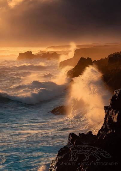 A dramatic seascape captures the rugged cliffs in Mornington as powerful waves crash against the dark rock. Golden light from the setting sun illuminates the sea spray, casting a warm glow over the turbulent ocean. The sky is brooding, with dense clouds parting slightly to let beams of sunlight streak across the churning waters and jagged coastline, creating a moody yet awe-inspiring atmosphere.