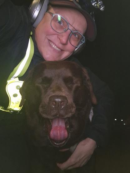Night time selfie of a person and a chocolate brown Labrador with greying snout. The person is wearing a black jacket, yellow safety harness, red cap, silver headphones and blue-rimmed glasses. On their cap rests a headstrap torch in off position. Their free hand is around the dog's shoulders. Both are facing the camera. The dog's mouth is wide open, as she's yawning wide! Taking selfies can be boring, I guess...