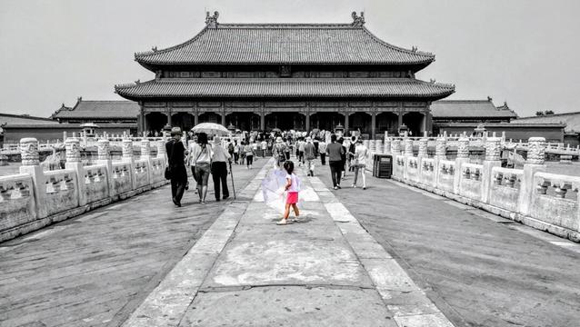 The image, primarily in black and white with selective colour, shows a grand view of the Hall of Supreme Harmony within the Forbidden City in Beijing, China. A grey stone pathway leads the viewer's eye toward the hall, flanked by ornate white stone railings. A group of people, rendered in monochrome, are walking along the pathway toward the hall. Among them, a young girl in a pink skirt stands out as the only element in colour. She holds an open umbrella pointed in front of her. The Hall of Supreme Harmony, a large traditional Chinese building with a multi-tiered roof, dominates the background. The sky is overcast, contributing to the muted tones of the scene.