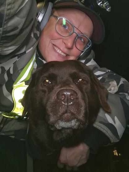 Night time selfie of a person and a chocolate brown Labrador with greying snout. The person is wearing a white-grey military camouflage jacket, yellow safety harness, red cap, silver headphones and blue-rimmed glasses. On their cap rests a headstrap torch in off position. Their free hand is around the dog's shoulders. Both are facing the camera.