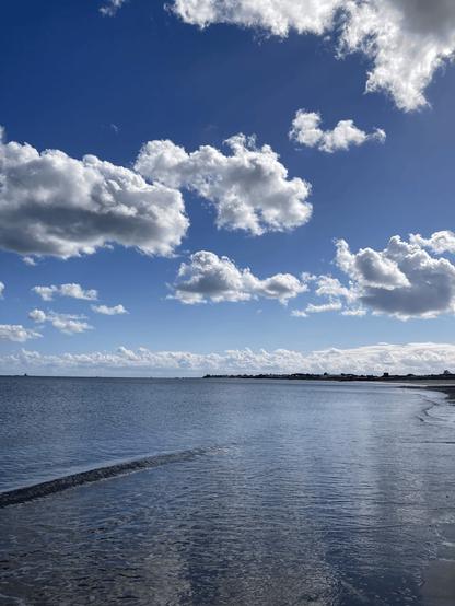 photo of the shore by the beach, with a reflection of the large blue sky above with clouds and rays of sunshine