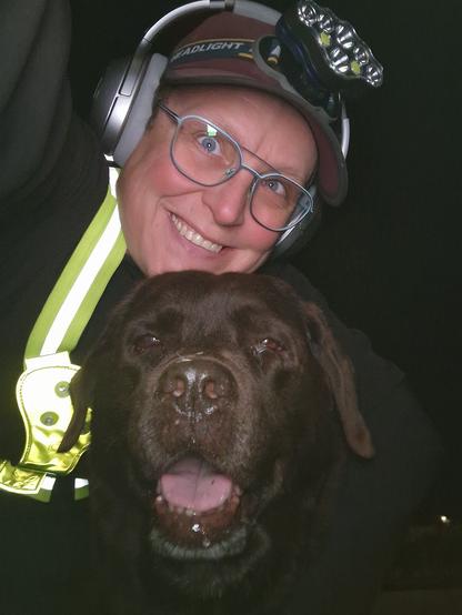 Night time selfie of a person and a chocolate brown Labrador with greying snout. The person is wearing a black jacket, yellow safety harness, red cap, silver headphones and blue-rimmed glasses. On their cap rests a headstrap torch in off position. Their free hand is around the dog's shoulders. Both are facing the camera. The dog's mouth is slightly open, showing off some blep. The person is smiling, enjoying their time spent together.