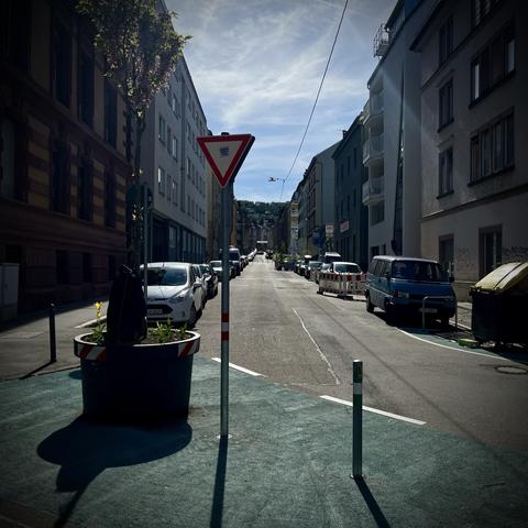 Foto auf der Augustenstraßen. Auf den Kreuzungen sind Diagonalsperren als modale Filter installiert, am Fahrbahnrand sind viele Autos geparkt und ein Schild „Vorfahrt achten“ signalisiert dem durch die Filter geleiteten Radverkehr, dass der Autoverkehr, der in U-Form in und wieder heraus aus der Augustenstraße geleitet wird, Vorfahrt hat. Auf der Kreuzung im modalen Filter steht ein junger Baum mit großem Blumentopf.