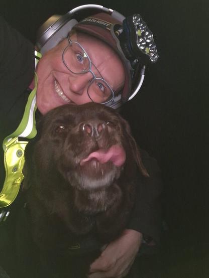 Night time selfie of a person and a chocolate brown Labrador with greying snout. The person is wearing a black jacket, yellow safety harness, red cap, silver headphones and blue-rimmed glasses. On their cap rests a headstrap torch in off position. Their free hand is around the dog's shoulders. Both are facing the camera. The dog's tongue is sticking out partially, as she was doing a mlem. The person is smiling, loving their time spent together.