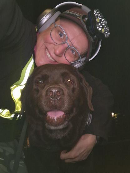 Night time selfie of a person and a chocolate brown Labrador with greying snout. The person is wearing a black jacket, yellow safety harness, red cap, silver headphones and blue-rimmed glasses. On their cap rests a headstrap torch in off position. Their free hand is around the dog's shoulders. Both are facing the camera. The dog's mouth is slightly open, showing off some mlem. The person's head is resting on the dog's head. They're smiling, enjoying every moment spent together.