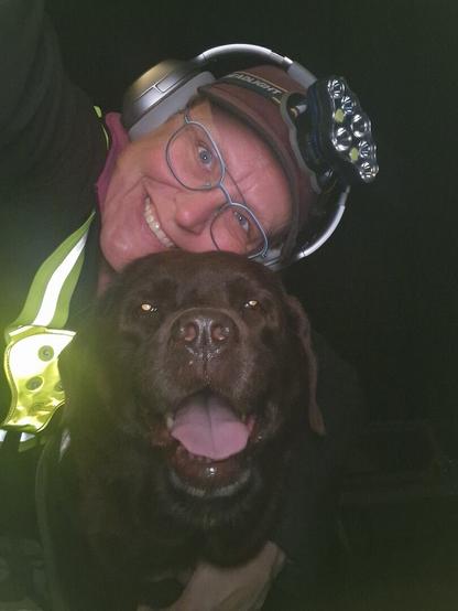 Night time selfie of a person and a chocolate brown Labrador with greying snout. The person is wearing a black jacket, yellow safety harness, red cap, silver headphones and blue-rimmed glasses. On their cap rests a headstrap torch in off position. Their free hand is around the dog's shoulders. Both are facing the camera. The dog's mouth is slightly open, showing off some blep. The person's head is resting on the dog's head. She's smiling, grateful for this time spent together.