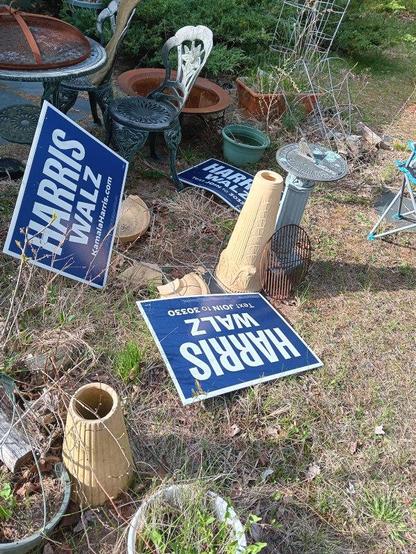A yard that is not well-tended. The grass is brown and uncut since the last season. Pieces of a broken ceramic birdbath, a metal grill that belongs in a barbecue, an off-balance sundial, and some rusty parts of different grill are scattered about. Three cardboard yard signs for Harris/Walz, the US Presidential campaign that lost to Trump, lay scattered across the yard as well.