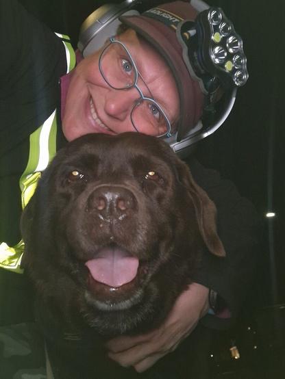 Night time selfie of a person and a chocolate brown Labrador with greying snout. The person is wearing a black jacket, yellow safety harness, red cap, silver headphones and blue-rimmed glasses. On their cap rests a headstrap torch in off position. Their free hand is around the dog's shoulders. Both are facing the camera. The dog's mouth is slightly open, showing off some blep. The person's head is resting on the dog's head. They're smiling, as they're grateful for the time spent together.