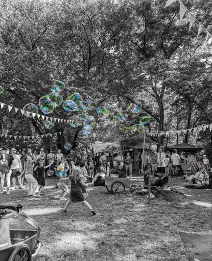 This is a black and white photograph of an outdoor event. A large number of soap bubbles are floating in the air, with their iridescent surfaces reflecting light. The bubbles are the only coloured part of the image. People of various ages are in the background, some looking at the bubbles. Bunting is strung between the trees, and there are tents and other structures in the background, suggesting a fair or festival. The overall scene is lively and festive.