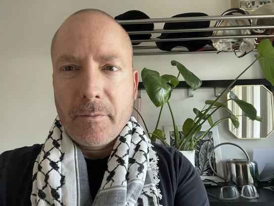 Me (white passing guy with short hair and dirty beard) standing in our hallway wearing my keffiyeh (traditional Palestinian white scarf with black motif and grey shading in parts) over a black t-shirt, in front of a plant, small mirror, some caps on a shelf, a square black abd white illustration of leaves, a metal watering can and a pair of glasses.