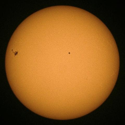 A bright sun with a uniform orange surface, featuring a large sunspot region visible on the left side and a smaller just off from center.