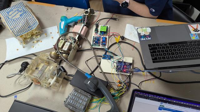 The keypad, ringer, and handset of a payphone are strewn about on a table with a multitude of wires connecting various parts to other electronics including a relay and an Arduino microcontroller.