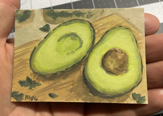 Miniature painting, held in the palm of a hand, of an avocado cut in half laying on a wood cutting board with a light background.