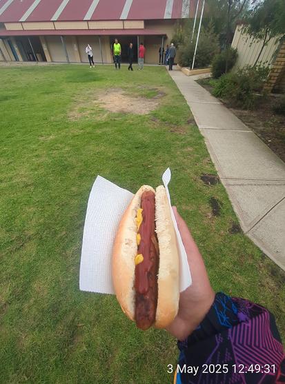 Picture with a democracy sausage held in a hand in the foreground, while in the background shows a voting location with no queue.