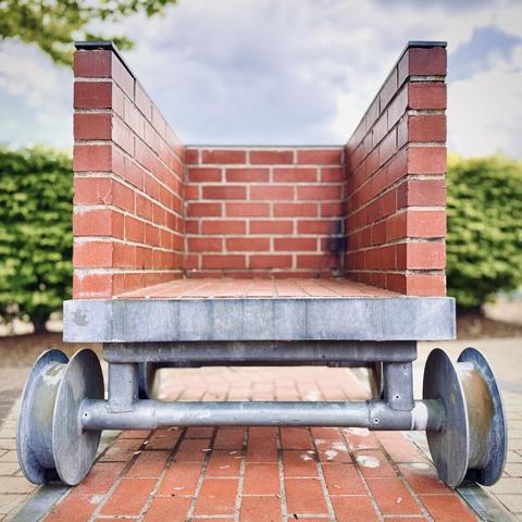 A red-brick, single-person train station bench, shaped like a stubby train car with the roof and one end removed, rests on fake train wheels cemented to fake train rails flush with the walkway on a mostly cloudy day.