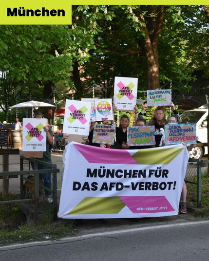 Das Foto zeigt eine Gruppe von 8-10 Menschen hinter einem Banner "München für das AfD-Verbot!". Die Menschen stehen auf einer Verkehrsinsel. Im Hintergrund ist ein Verkaufsstand unter Bäumen zu sehen. Die Menschen halten Schilder in die Kamera auf denen steht: "AfD-Verbot Jetzt", "Kein Platz für Nazis", "Erika Mustermann will das AfD-Verbot", "Max Mustermann auch!", "Kathrin Mustermann-Huber auch!" und "Mustermanns Lump auch!"