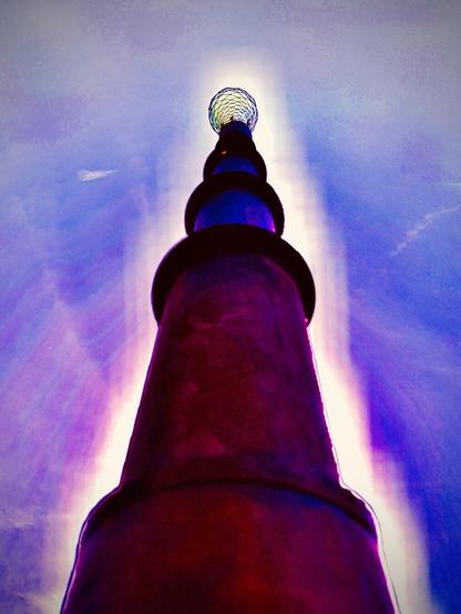 A metallic, sectioned light post-like sculpture towers into the sky, the colors enhanced and shifted to red, purple, and blue extremes.