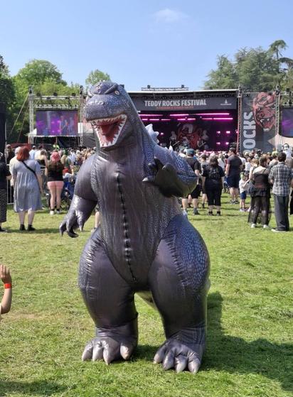 A large inflatable Godzilla stands in front of a large festival stage in the daytime. It is sunny and there is a crowd of people behind.