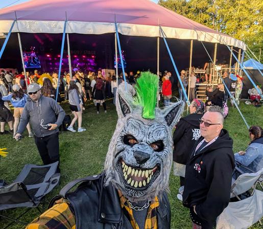 A grey werewolf stands in front of a festival big top stage with a crowd inside - he has a green mohawk, red/yellow eyes and is wearing a yellow check shirt and a leather vest.