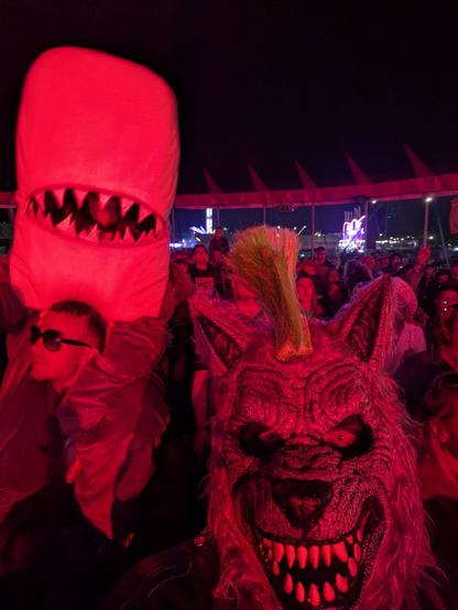A grey werewolf stands in festival big top with a crowd inside - the scene is lit in red light from the stage - he has a green mohawk. There is a child in a shark costume sitting on a mans shoulders behind.