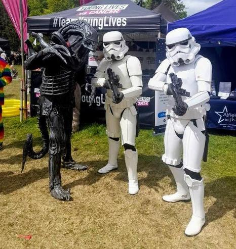 A black Xenomorph Alien stands with two Star Wars Stormtroopers in front of a charity gazebo, on a grass surface outside.