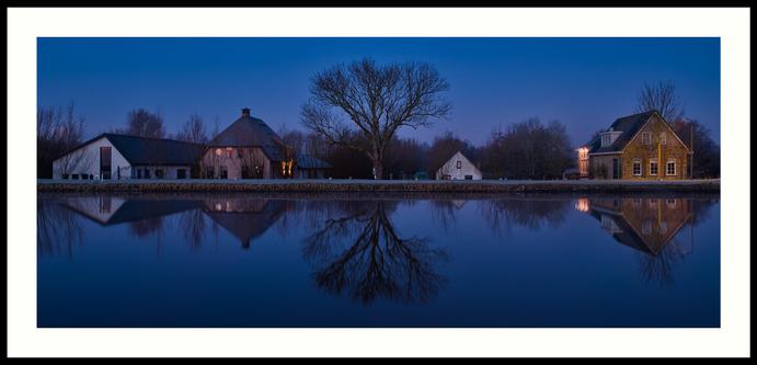 colour - Blue hour photo of part of the waterfront along the Delftse Schie. In the middle of this shot stands a, still leafless, large oak tree with two plots with houses and barns to the left and right of it, creating a vertical symmetry. Due to the late blue hour, everything has a somewhat blueish colour. The house on the right is warmly lit by a single street lamp and, reflected in the windows of the house on the left, the first warm tones of dawn are visible. The upper part of this scene is reflected in the water, creating a horizontal symmetry.