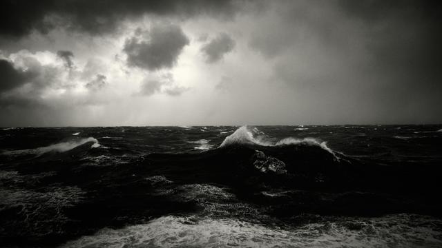 The image shows a tumultuous ocean with dark, rolling waves under a cloudy sky. The waves are high and the water appears choppy. The clouds are heavy and dark, indicating an approaching storm. The scene conveys a sense of intensity and power.