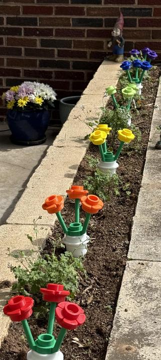 A planter box featuring colorful artificial flowers made from plastic, with flowers in shades of red, orange, yellow, green, and blue. A small gnome figurine stands in the background next to potted plants.
