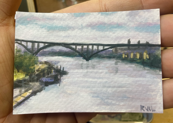 Miniature painting, held in the palm of a hand, of a very high arch bridge over a river with a bright winter sky reflected in the water.