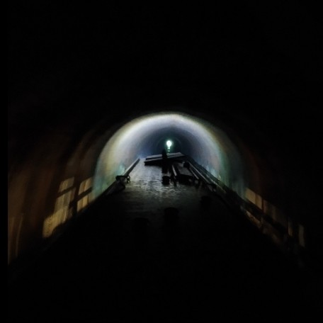 A narrowboat in a dark canal tunnel, which has a circular cross-section over the water, walls and curved "ceiling", the lower half filled with water. A light on the bow illuminates a small section ahead, an arc of light on tunnel walls and ceiling. In the distance is the end of the tunnel, a speck of daylight the size of a coin when held at arm's length. Lights inside the boat also illuminate the tunnel walls, but faintly, as rectangular projections of the window openings in either side of the steel narrowboat. On the roof of the boat there's some equipment, unrecognisable in the dark: a barge pole (the proverbial "ten-foot pole"), a hook, a plank, and two solar panels.