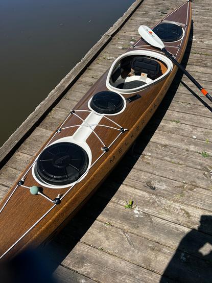 Photograph of a kayak on a dock. The kayak is light brown in color, made of cork and flax. There are three hatches, two in front of the cockpit and one aft, and the areas surrounding the hatches are bright white. The cockpit is shaped like a guitar pick. A white-bladed paddle rests on the back of the kayak.
