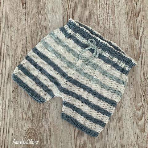 Kurze gestrickte Hose für Babys. Die Hose hat weiße, blaugraue und grünblaue Streifen und ist mit einer Kordel am Bündchen zu schließen.