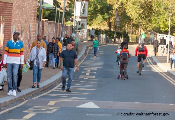 A narrow urban street has school zigzag markings down one side, there are adults and children walking, scooting and cycling along the whole street space. No cars are present.