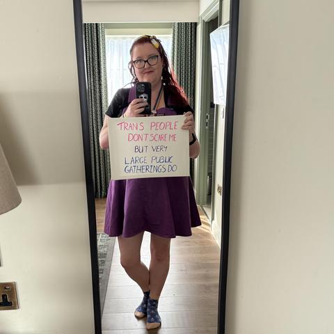 A mirror selfie of a white person with red hair, pushed back by a bandana the colour of the nonbinary pride flag. They are wearing a black tshirt and a purple dungaree dress, holding a sign saying Trans People Don’t Scare Me But Large Public Gatherings Do’ over their chest.