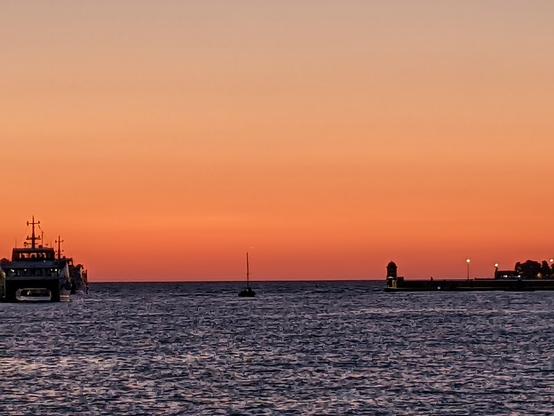 The image depicts a serene seascape at sunset. The sky is painted with a gradient of warm colors, transitioning from a soft peach near the horizon to a deeper orange higher up. The calm sea reflects these hues, creating a tranquil atmosphere. On the left side of the image, a large boat with multiple masts is visible, silhouetted against the vibrant sky. In the center, a smaller boat with a single mast floats on the water. On the right, a lighthouse stands on a pier, with a few lights turned on, adding a touch of human presence to the natural scene. The overall composition emphasizes the beauty of the sunset and the peacefulness of the maritime environment.

Provided by @altbot