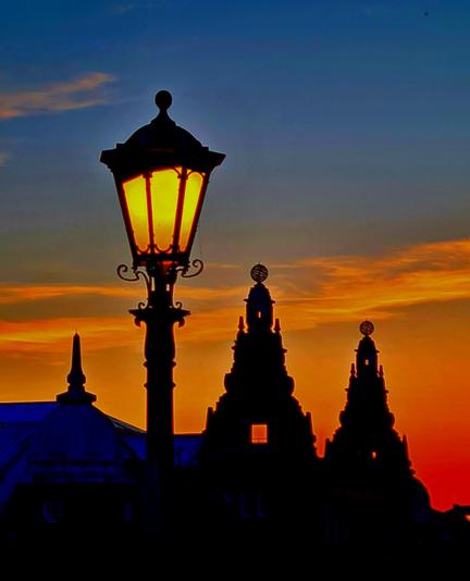 An old gas lamp, now lit by electricity, and the ornate finials of a 1920s exhibition hall against an orange sunset.