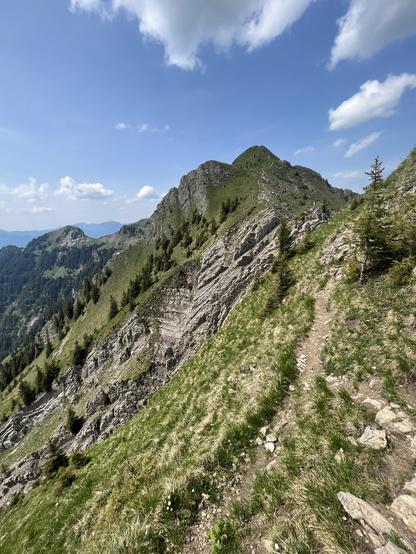 Foto einer Berglandschaft. Im Vordergrund ein schmaler Bergwanderweg entlang eines Abhangs.