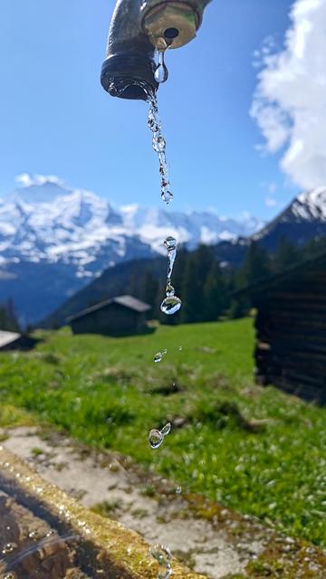 A close-up shot captures crystal-clear water droplets falling from a mountain tap, frozen mid-air in perfect clarity. In the blurred background, a vivid alpine scene unfolds — lush green meadows, rustic wooden huts, and towering snow-covered peaks under a bright blue sky. The image beautifully contrasts the fine detail of the droplets with the grandeur of the mountains beyond, evoking freshness, purity, and the quiet magic of high-altitude life.