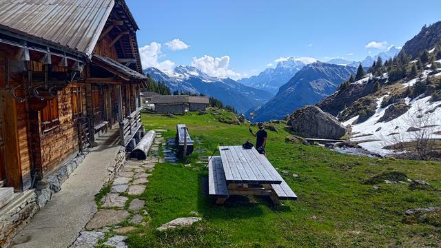 A rustic alpine hut with wooden shutters and a stone foundation sits quietly on a sunlit slope, surrounded by vivid green grass and melting snowfields. A wooden bench and picnic table offer a peaceful resting spot, while a hiker stands nearby, capturing the moment against a breathtaking backdrop of jagged, snow-covered peaks and a deep blue sky. A narrow stream trickles past a large boulder, adding to the serene mountain atmosphere.