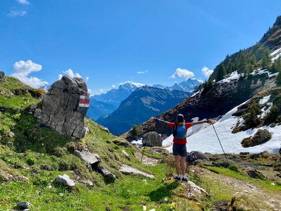 A hiker stands on a scenic alpine trail, gazing toward a majestic, snow-capped mountain range under a clear blue sky. The path winds past a marked trail stone, patches of melting snow, and spring greenery — a perfect snapshot of high-altitude hiking in the Swiss Alps.